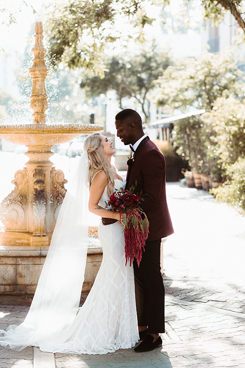 groom in a burgundy tuxedo and black bow tie and the bride in a white formfitting gown with thin straps and cathedral length veil