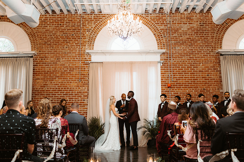 bride in a formfitting lace white gown with thin straps, and the groom in a burgundy tuxedo with a black bow tie.