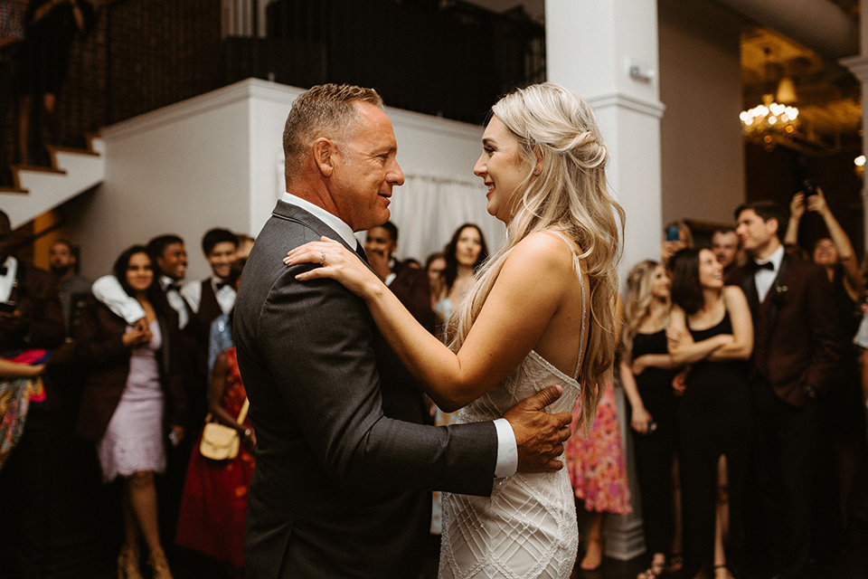 bride in a white formfitting gown with thin straps and her father in a dark grey suit