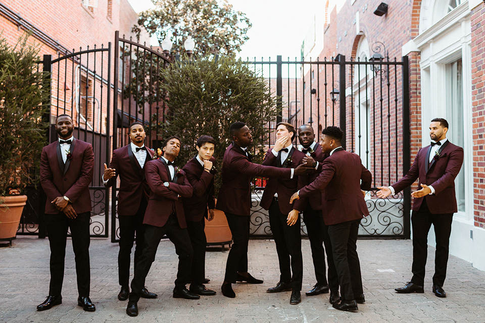  the groom in a burgundy tuxedo with a black bow tie and the groomsmen in burgundy tuxedos