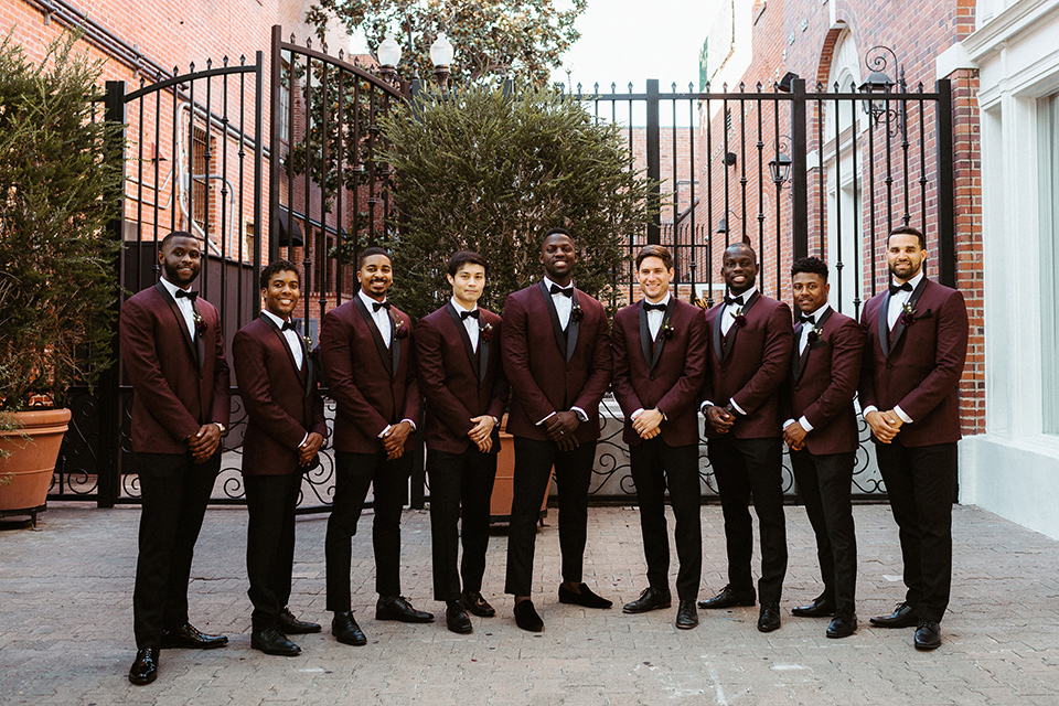  the groom in a burgundy tuxedo with a black bow tie and the groomsmen in burgundy tuxedos