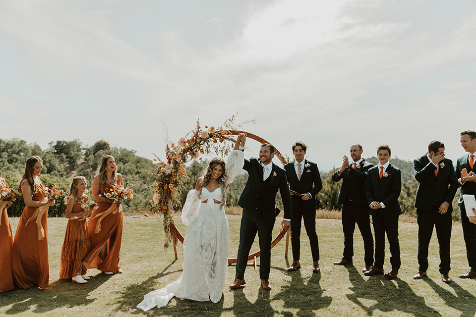  the bridesmaids in deep orange gowns, the groomsmen in navy suits, and the bride in an ivory rue de sine gown and the groom in a dark blue suit with an orange tie at the ceremony