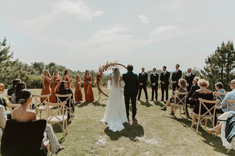  the bridesmaids in deep orange gowns, the groomsmen in navy suits, and the bride in an ivory rue de sine gown and the groom in a dark blue suit with an orange tie at the ceremony