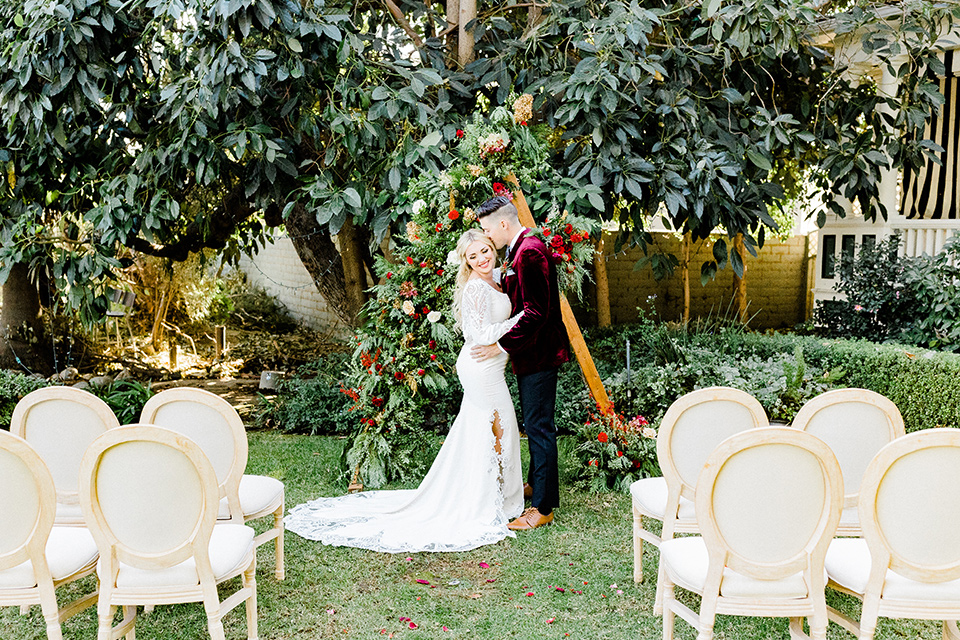  Christmas styled shoot with the groom in a burgundy velvet tuxedo and black pants  and the bride in a white long formfitting gown and long sleeves 