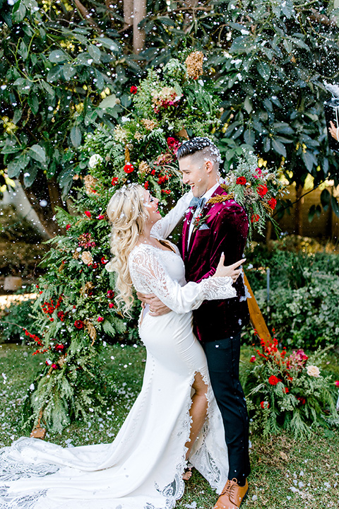  Christmas styled shoot with the groom in a burgundy velvet tuxedo and black pants  and the bride in a white long formfitting gown and long sleeves 