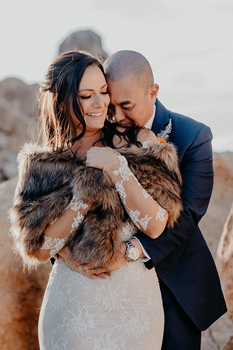  bride in a formfitting gown and a faux fur shawl and the groom in a dark blue notch lapel suit 