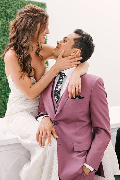  bride in a fashionable white jump suit with a strapless neckline and wide leg and the groom in a rose pink suit with a black floral neck tie