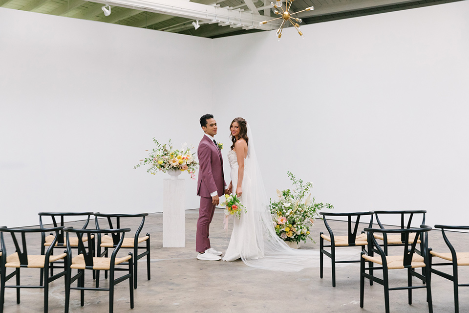  bride in a fashionable white jump suit with a strapless neckline and wide leg and the groom in a rose pink suit with a black floral neck tie 