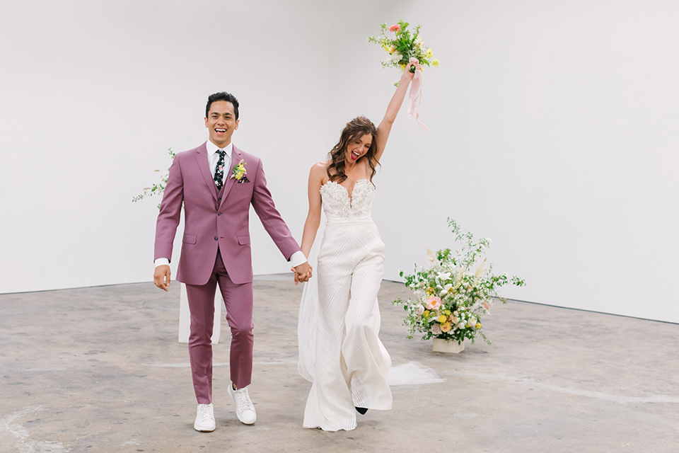  bride in a fashionable white jump suit with a strapless neckline and wide leg and the groom in a rose pink suit with a black floral neck tie 