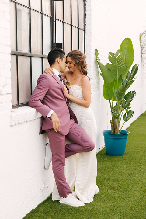  bride in a fashionable white jump suit with a strapless neckline and wide leg and the groom in a rose pink suit with a black floral neck tie