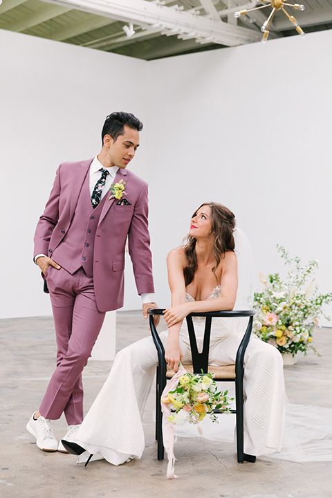  bride in a fashionable white jump suit with a strapless neckline and wide leg and the groom in a rose pink suit with a black floral neck tie