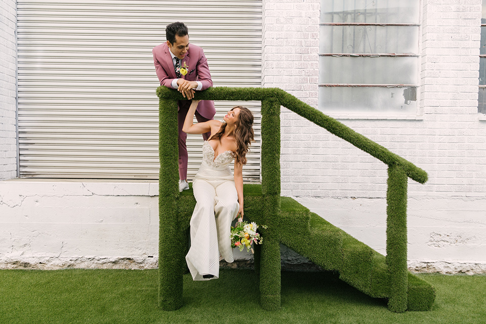  bride in a fashionable white jump suit with a strapless neckline and wide leg and the groom in a rose pink suit with a black floral neck tie 