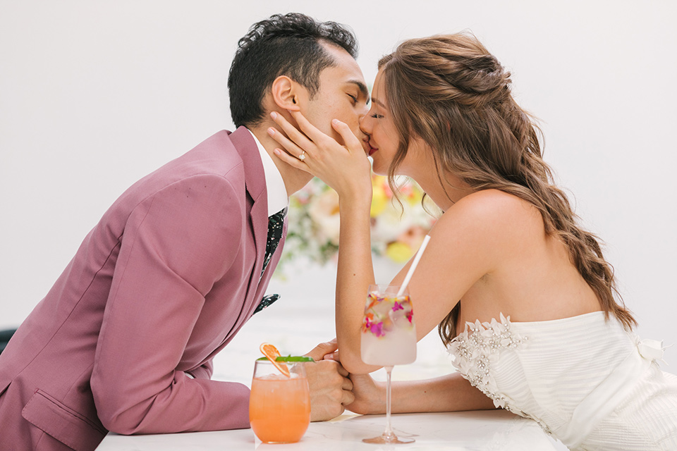  bride in a fashionable white jump suit with a strapless neckline and wide leg and the groom in a rose pink suit with a black floral neck tie 