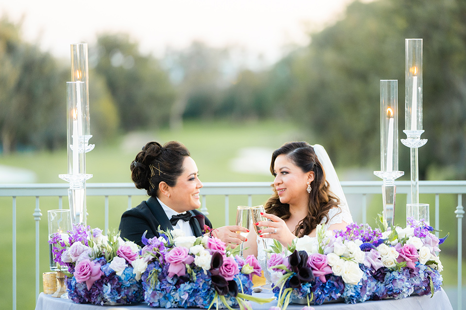  one bride in a black women’s tuxedo with a black diamond shaped bow tie and one bride in a white mermaid style gown with a beaded bodice and strapless neckline 