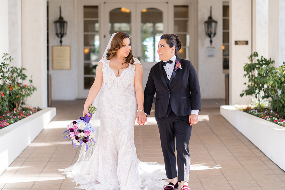  one bride in a black women’s tuxedo with a black diamond shaped bow tie and one bride in a white mermaid style gown with a beaded bodice and strapless neckline 