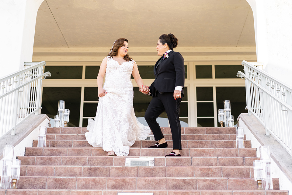  one bride in a black women’s tuxedo with a black diamond shaped bow tie and one bride in a white mermaid style gown with a beaded bodice and strapless neckline 