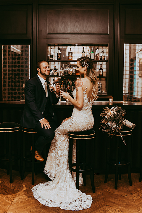  groom in a navy suit and bow tie and the bride in a white lace strapless gown sitting at the bar 
