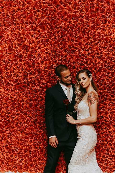  groom in a navy suit and bow tie and the bride in a white lace strapless gown in front of red wall 