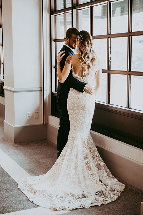  groom in a navy suit and bow tie and the bride in a white lace strapless gown looking out the window 