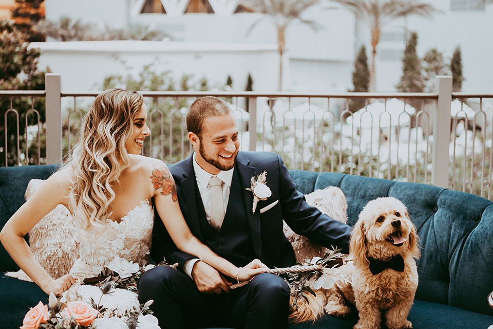  groom in a navy suit and bow tie and the bride in a white lace strapless gown holding their dog