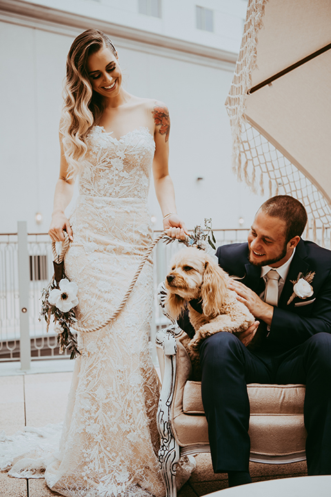  groom in a navy suit and bow tie and the bride in a white lace strapless gown with their dog