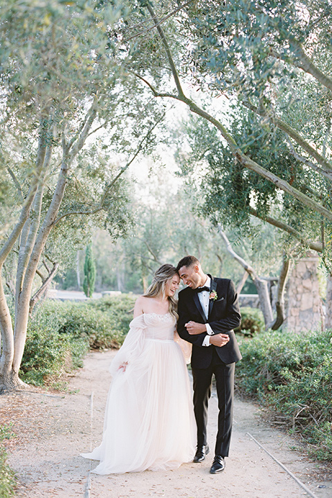  bride in a blush gown with off the shoulder sleeves and the groom in a black notch lapel tuxedo and black bow tie 