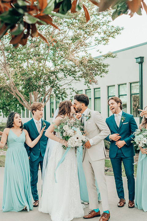  bride in a white gown and the groom in a tan notch lapel suit with a floral tie, the groomsmen in blue suits and the bridesmaids in light blue gowns 