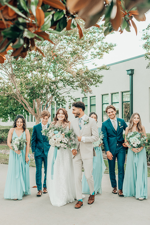  bride in a white gown and the groom in a tan notch lapel suit with a floral tie, the groomsmen in blue suits and the bridesmaids in light blue gowns 