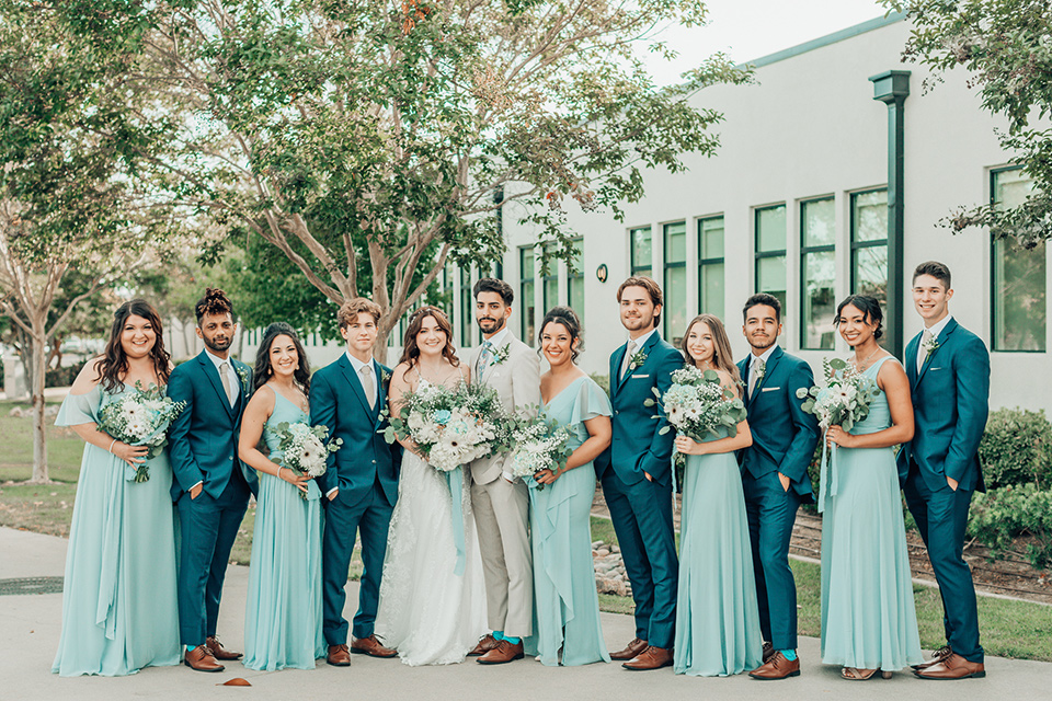  bride in a white gown and the groom in a tan notch lapel suit with a floral tie, the groomsmen in blue suits and the bridesmaids in light blue gowns 
