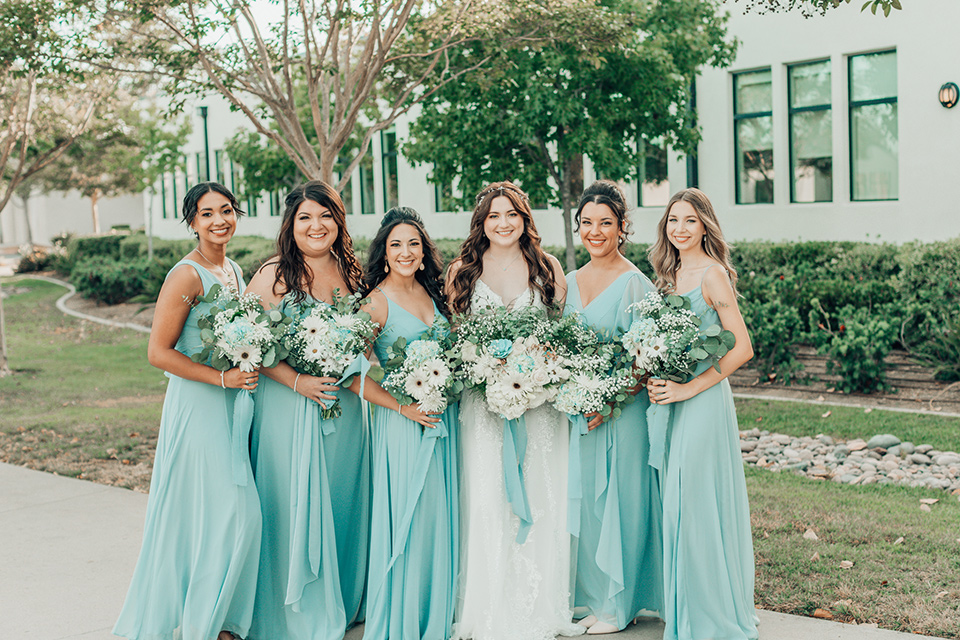  bride in a white gown and the groom in a tan notch lapel suit with a floral tie, the groomsmen in blue suits and the bridesmaids in light blue gowns 