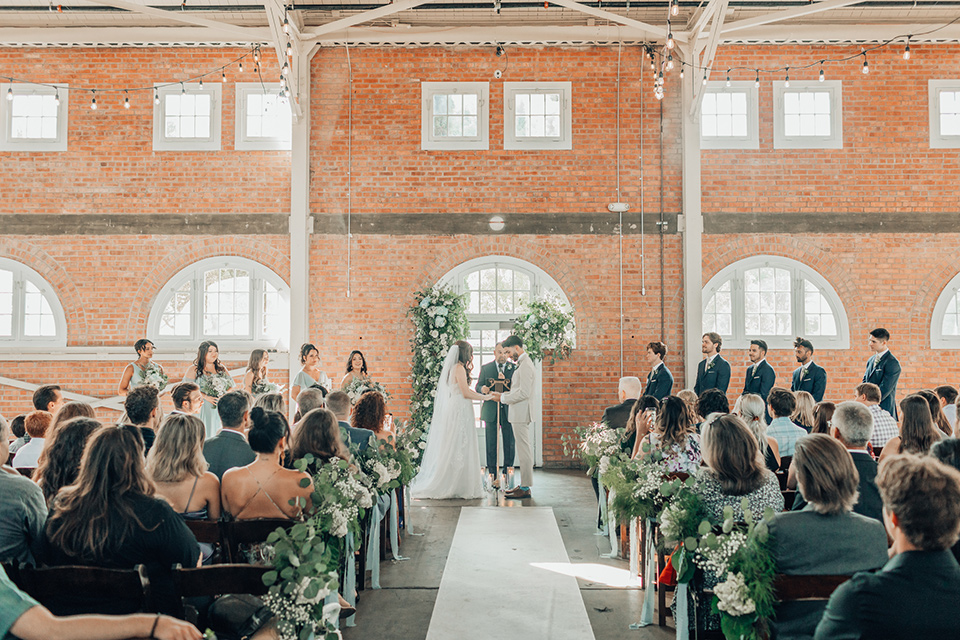  bride in a white gown and the groom in a tan notch lapel suit with a floral tie, the groomsmen in blue suits and the bridesmaids in light blue gowns 