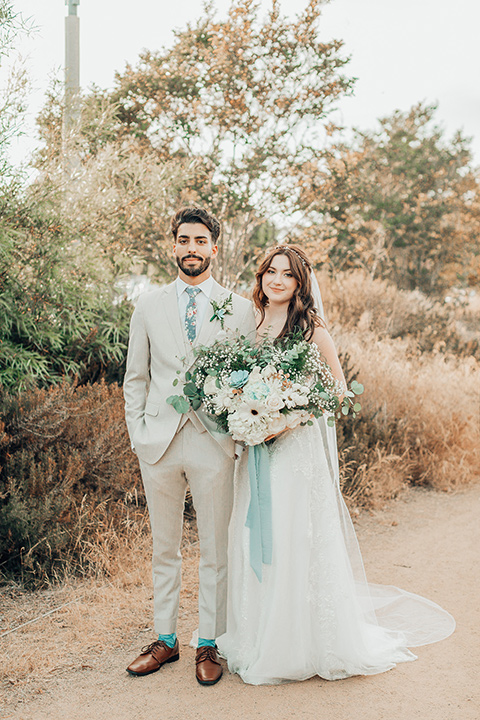  bride in a white gown and the groom in a tan notch lapel suit with a floral tie, the groomsmen in blue suits and the bridesmaids in light blue gowns 