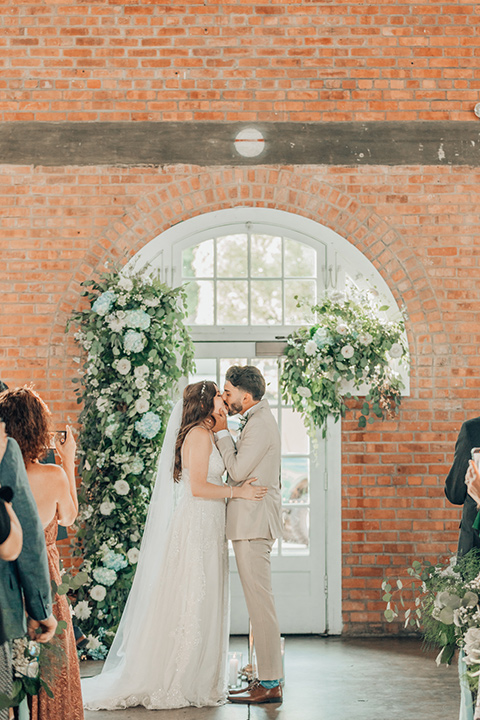  bride in a white gown and the groom in a tan notch lapel suit with a floral tie, the groomsmen in blue suits and the bridesmaids in light blue gowns 