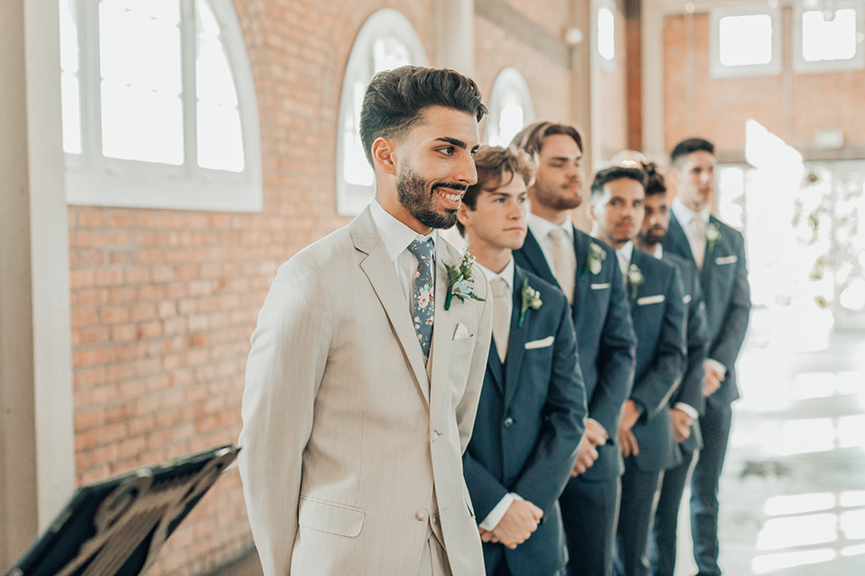  bride in a white gown and the groom in a tan notch lapel suit with a floral tie, the groomsmen in blue suits and the bridesmaids in light blue gowns 