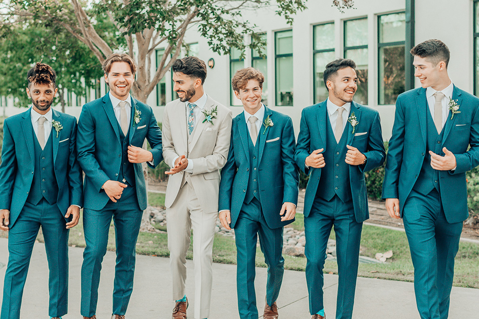  bride in a white gown and the groom in a tan notch lapel suit with a floral tie, the groomsmen in blue suits and the bridesmaids in light blue gowns 