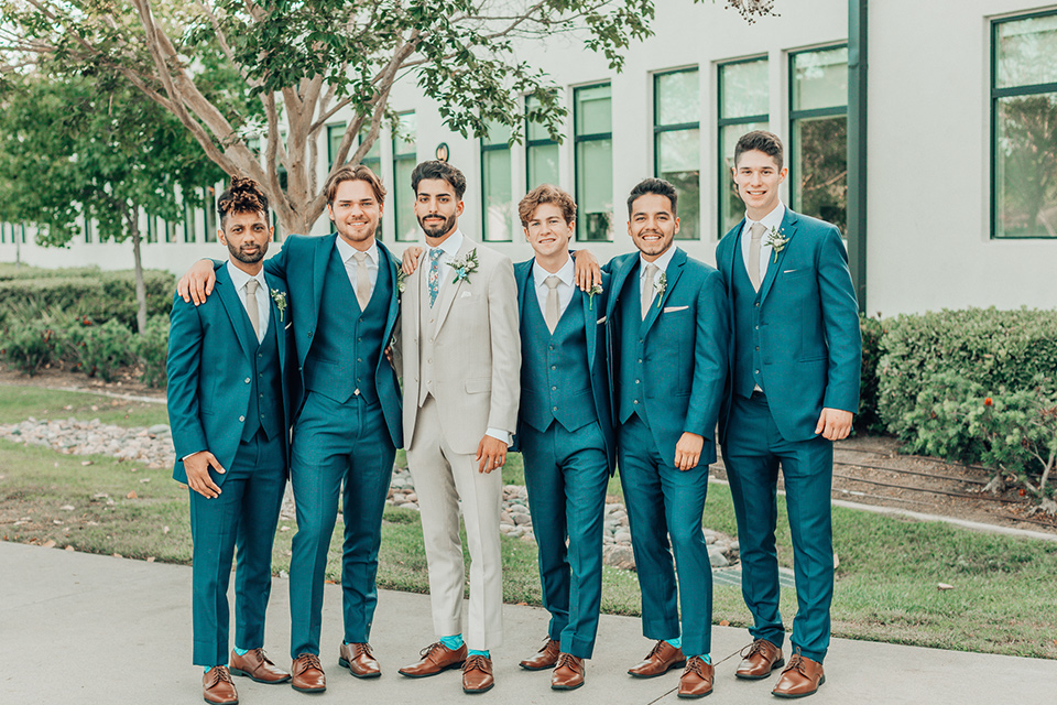  bride in a white gown and the groom in a tan notch lapel suit with a floral tie, the groomsmen in blue suits and the bridesmaids in light blue gowns 