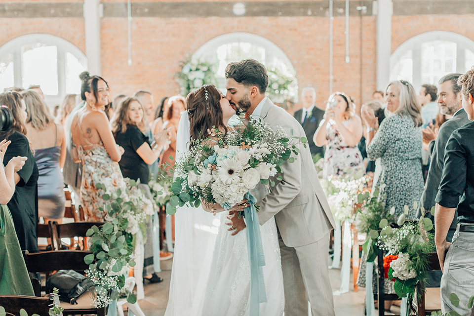  bride in a white gown and the groom in a tan notch lapel suit with a floral tie, the groomsmen in blue suits and the bridesmaids in light blue gowns 