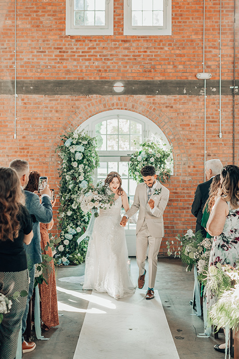  bride in a white gown and the groom in a tan notch lapel suit with a floral tie, the groomsmen in blue suits and the bridesmaids in light blue gowns 