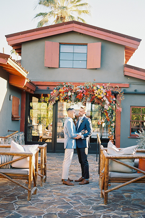  a groom in a blue notch lapel coat and charcoal grey pants and the other groom in a light blue coat and tan pants