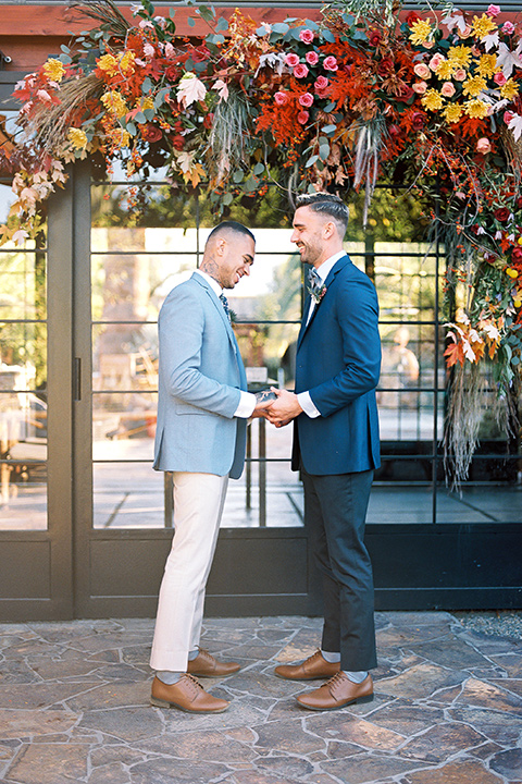  a groom in a blue notch lapel coat and charcoal grey pants and the other groom in a light blue coat and tan pants 