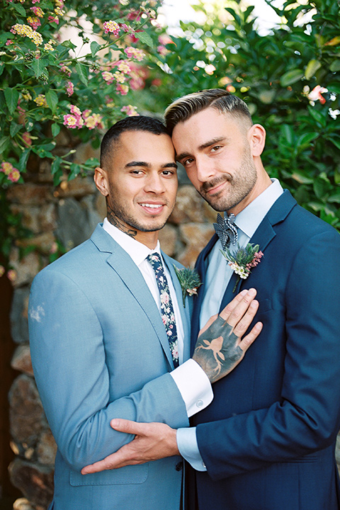  a groom in a blue notch lapel coat and charcoal grey pants and the other groom in a light blue coat and tan pants