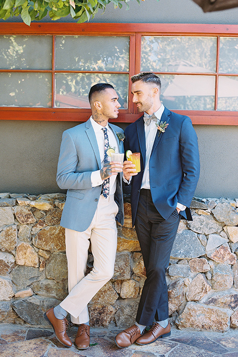  a groom in a blue notch lapel coat and charcoal grey pants and the other groom in a light blue coat and tan pants 