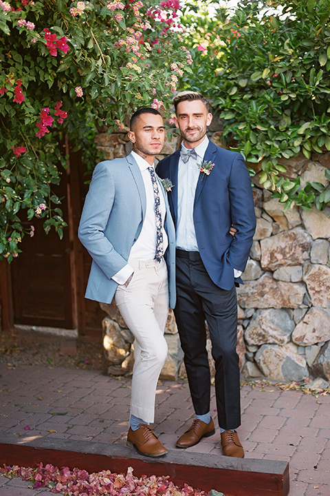  a groom in a blue notch lapel coat and charcoal grey pants and the other groom in a light blue coat and tan pants