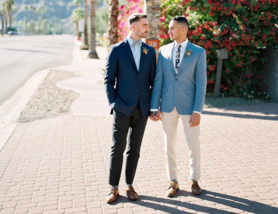  a groom in a blue notch lapel coat and charcoal grey pants and the other groom in a light blue coat and tan pants 