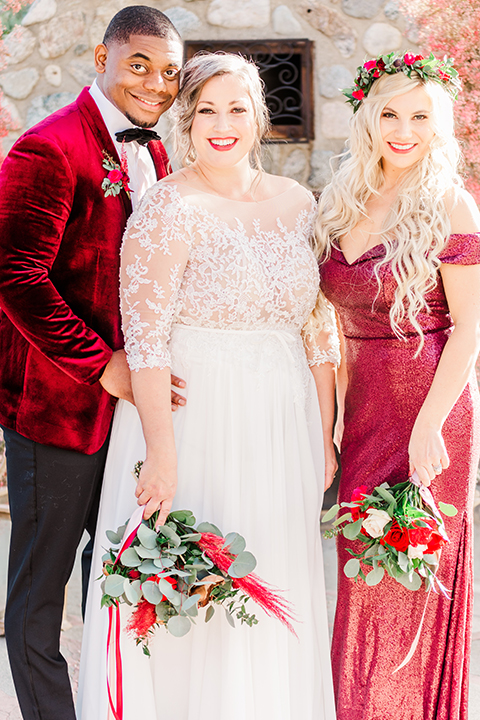  bride in a white and ivory gown with a natural waist and lace long sleeves, the groom in a burgundy velvet tuxedo with a black bow tie, and the bridesmaid in a red velvet long gown and a floral headpiece