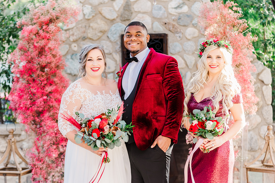  bride in a white and ivory gown with a natural waist and lace long sleeves, the groom in a burgundy velvet tuxedo with a black bow tie, and the bridesmaid in a red velvet long gown and a floral headpiece