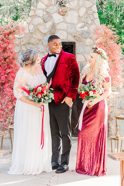  bride in a white and ivory gown with a natural waist and lace long sleeves, the groom in a burgundy velvet tuxedo with a black bow tie, and the bridesmaid in a red velvet long gown and a floral headpiece