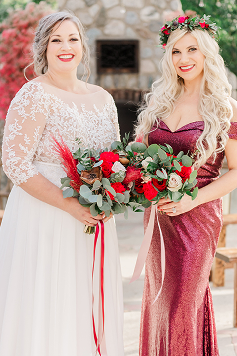  bride in a white and ivory gown with a natural waist and lace long sleeves and the bridesmaid in a red velvet long gown and a floral headpiece