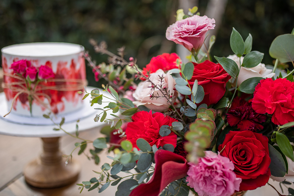  bride in a white and ivory gown with a natural waist and lace long sleeves, the groom in a burgundy velvet tuxedo with a black bow tie, and the bridesmaid in a red velvet long gown and a floral headpiece