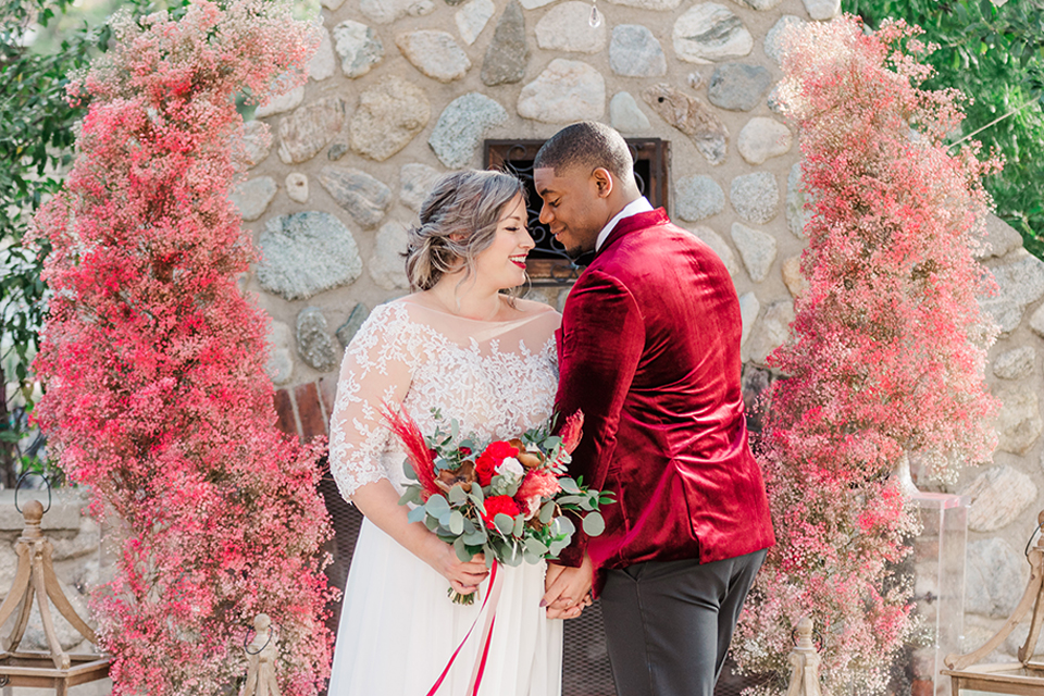  bride in a white and ivory gown with a natural waist and lace long sleeves, the groom in a burgundy velvet tuxedo with a black bow tie, and the bridesmaid in a red velvet long gown and a floral headpiece